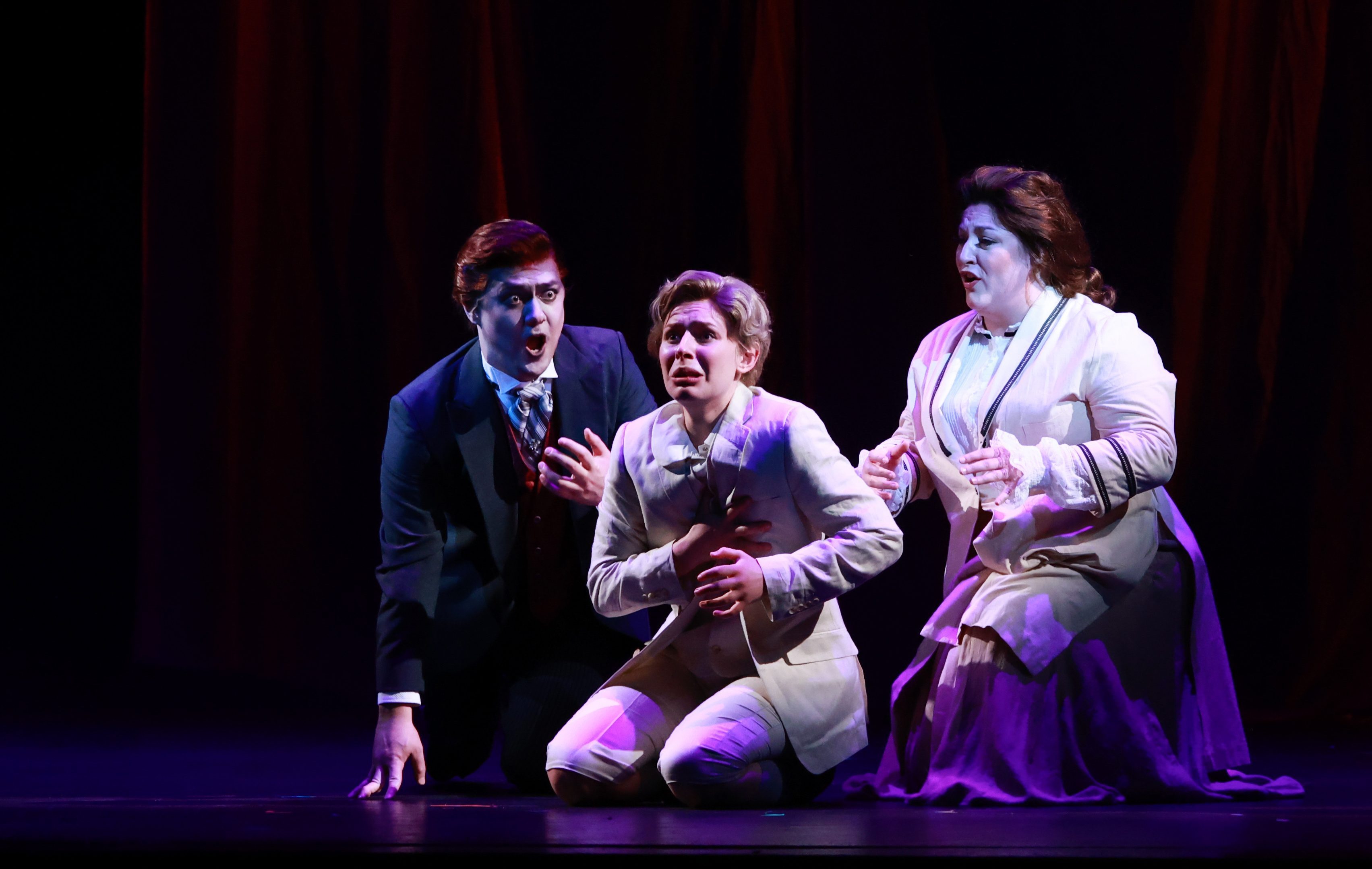 Three people dressed in 19th-century clothing on stage with a dark background. The man in the center kneels. The person in...